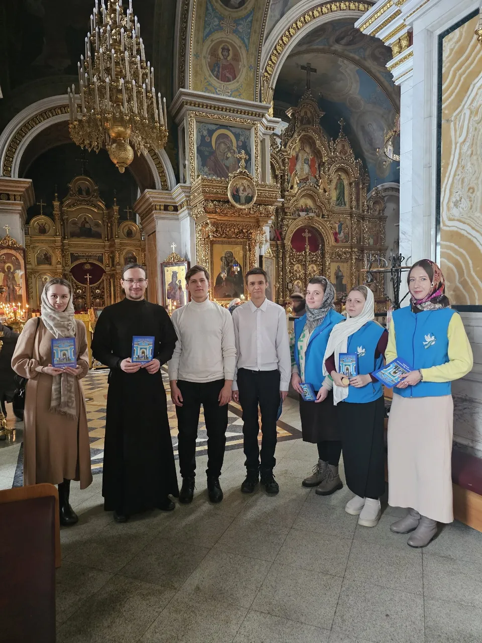Молодые волонтёры в синих жилетах с брошюрами стоят рядом со священником в храме